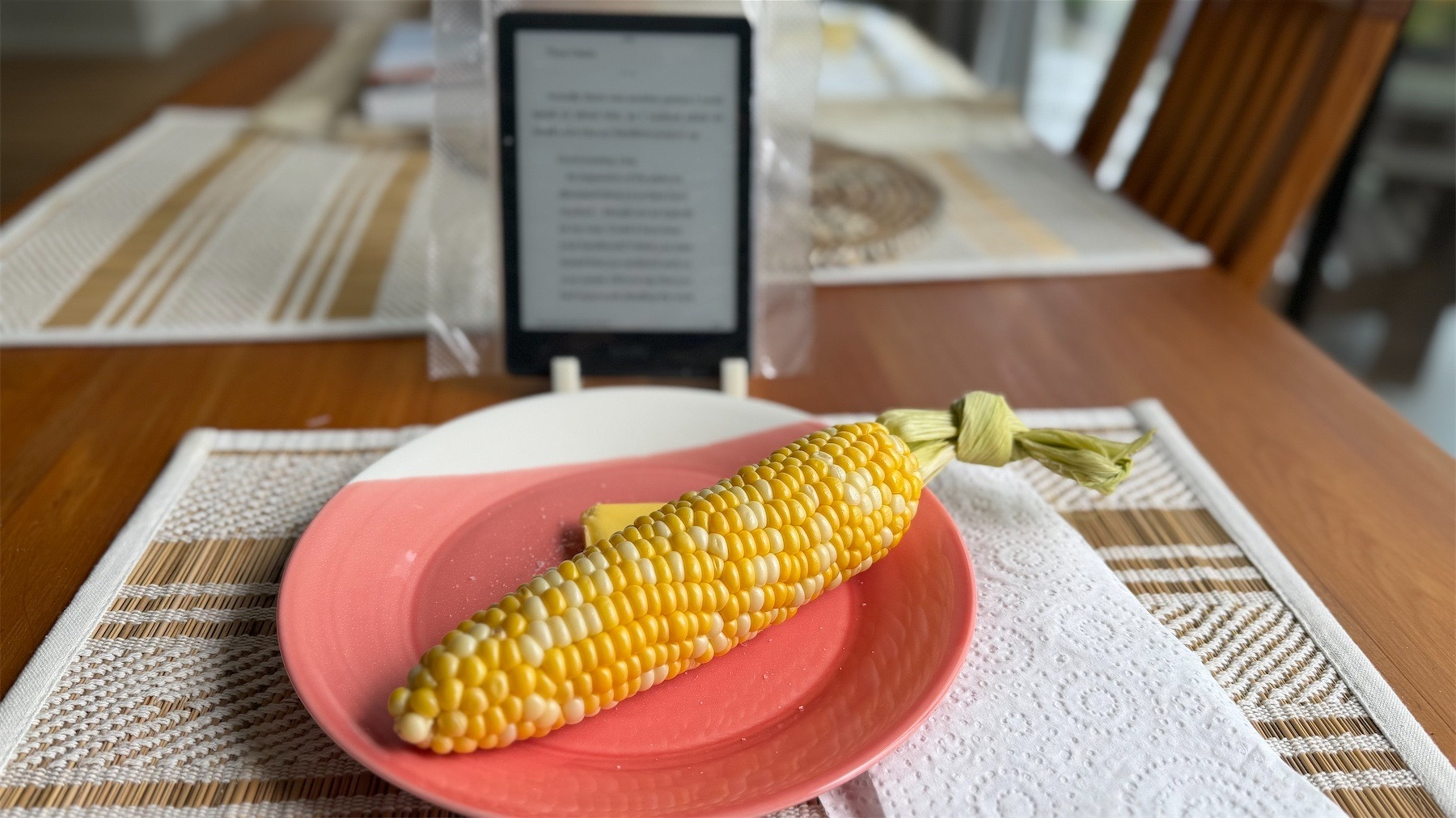 Table with plate with corn cob, behind which is a Kindle is a plastic cover.