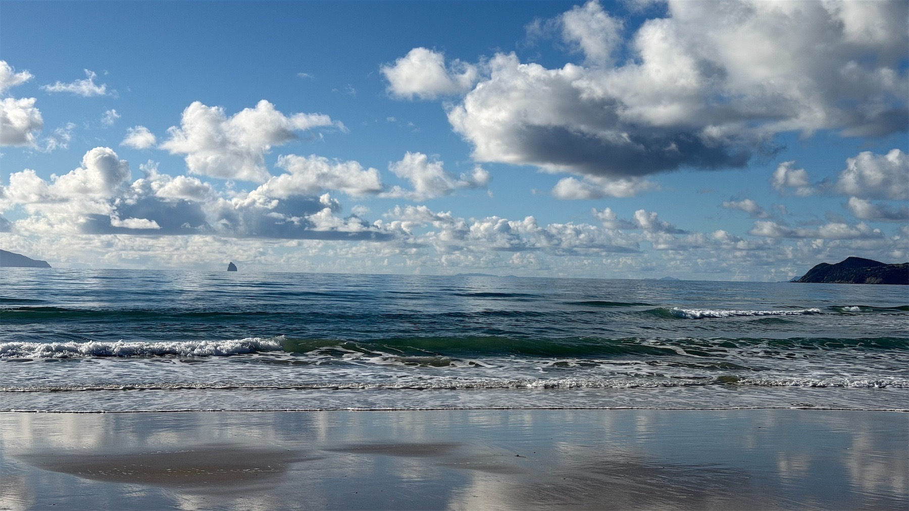 View from beach across the sea to some small islands, with large white and grey puffy clouds against a blue sky.