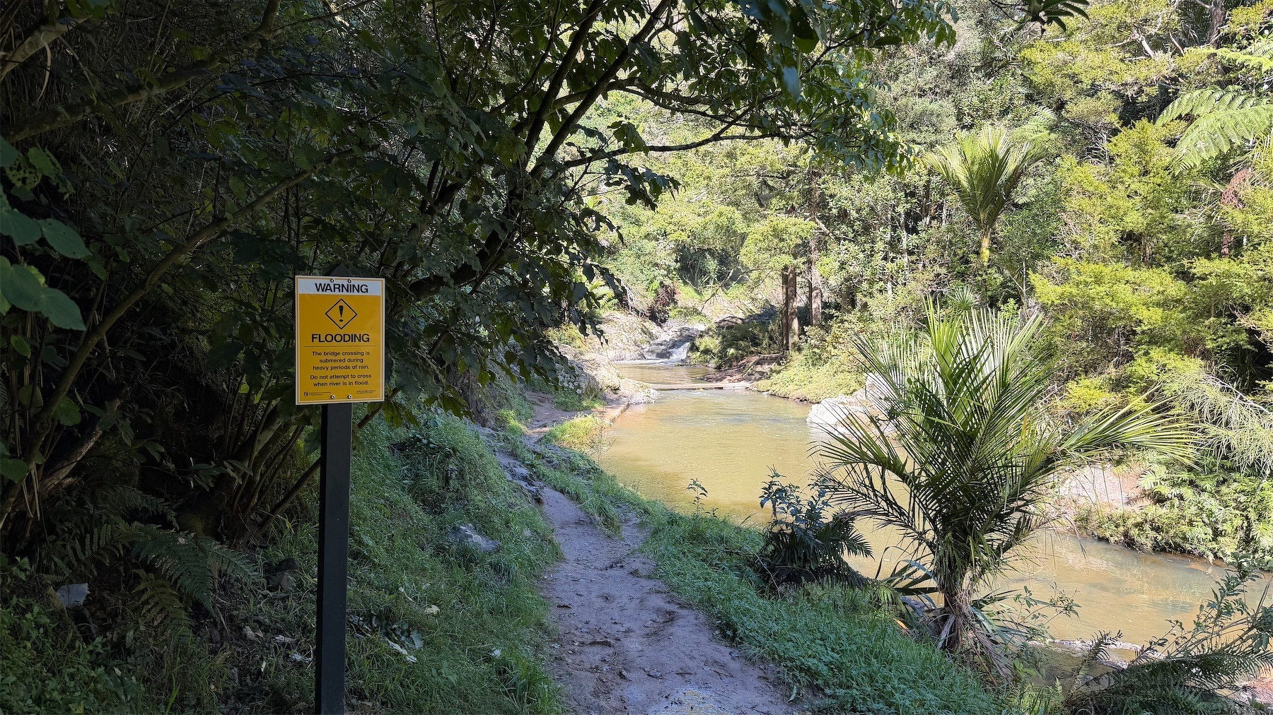 Sign warns of flooding risk. Narrow muddy path beside a stream in bush. 