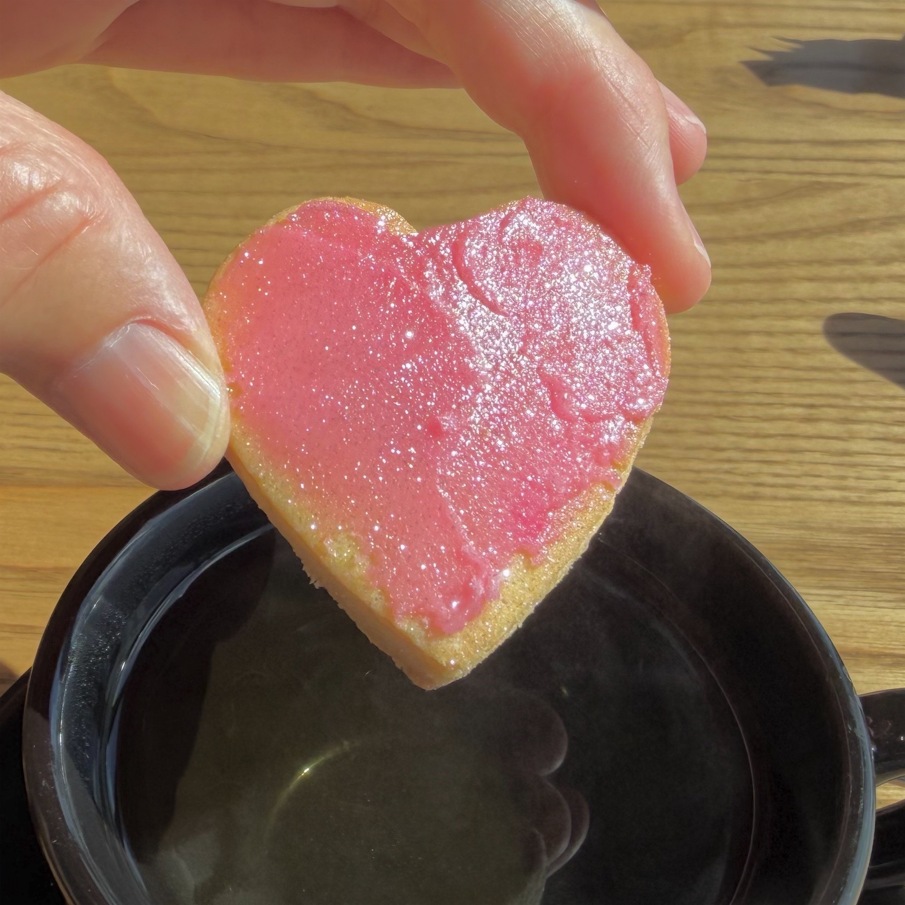 Fingers holding a shortbread heart with pink icing above a cup of tea.