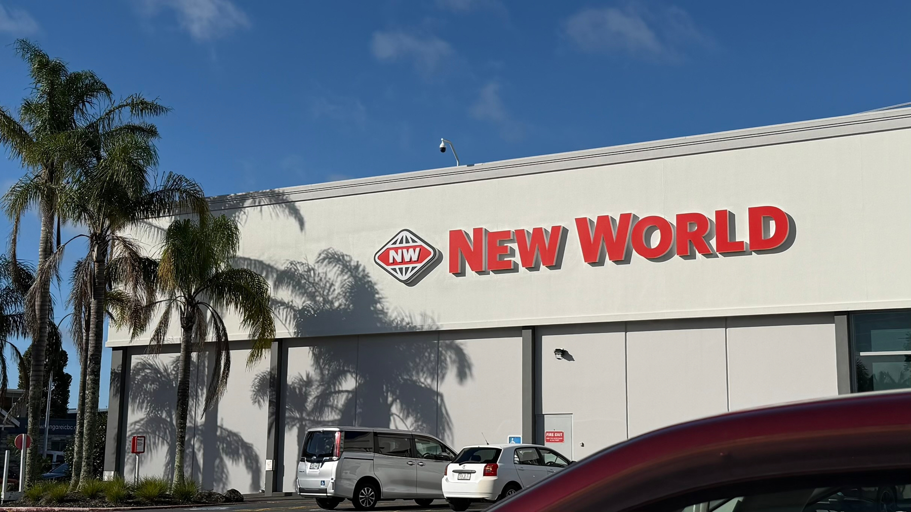 A New World supermarket building is shown with a few cars parked in front and palm trees nearby.