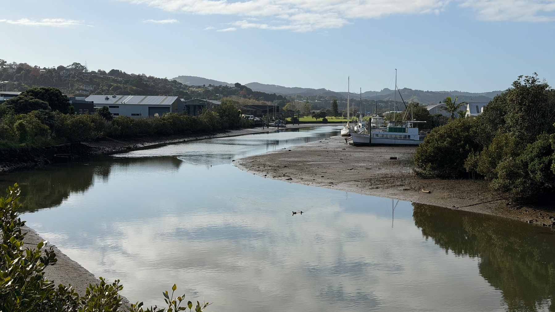 A calm river scene features boats moored alongside its banks, surrounded by lush greenery and hills under a partly cloudy sky.