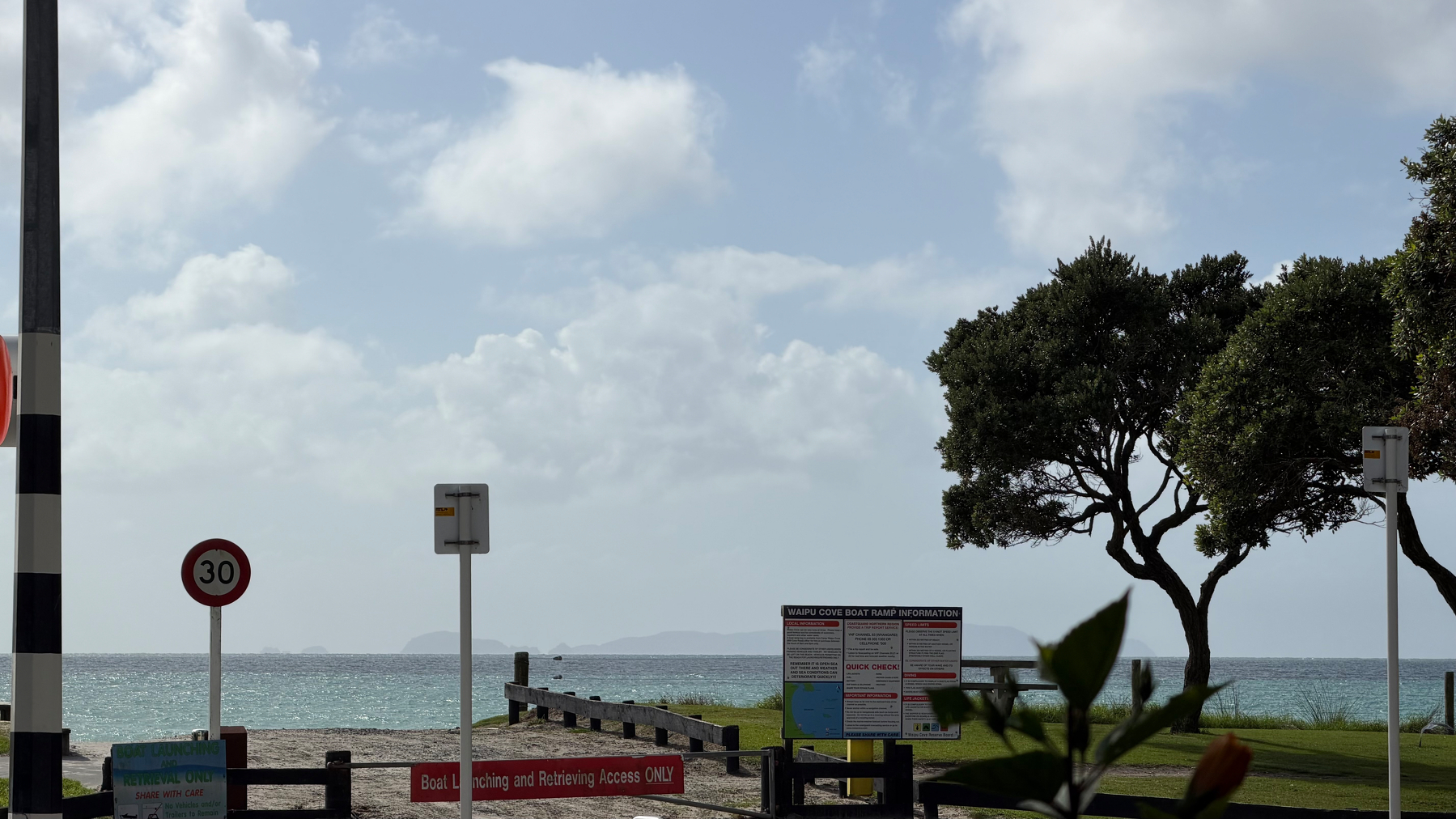 A serene coastal view features a pathway leading to the ocean, flanked by signs and trees under a partly cloudy sky.