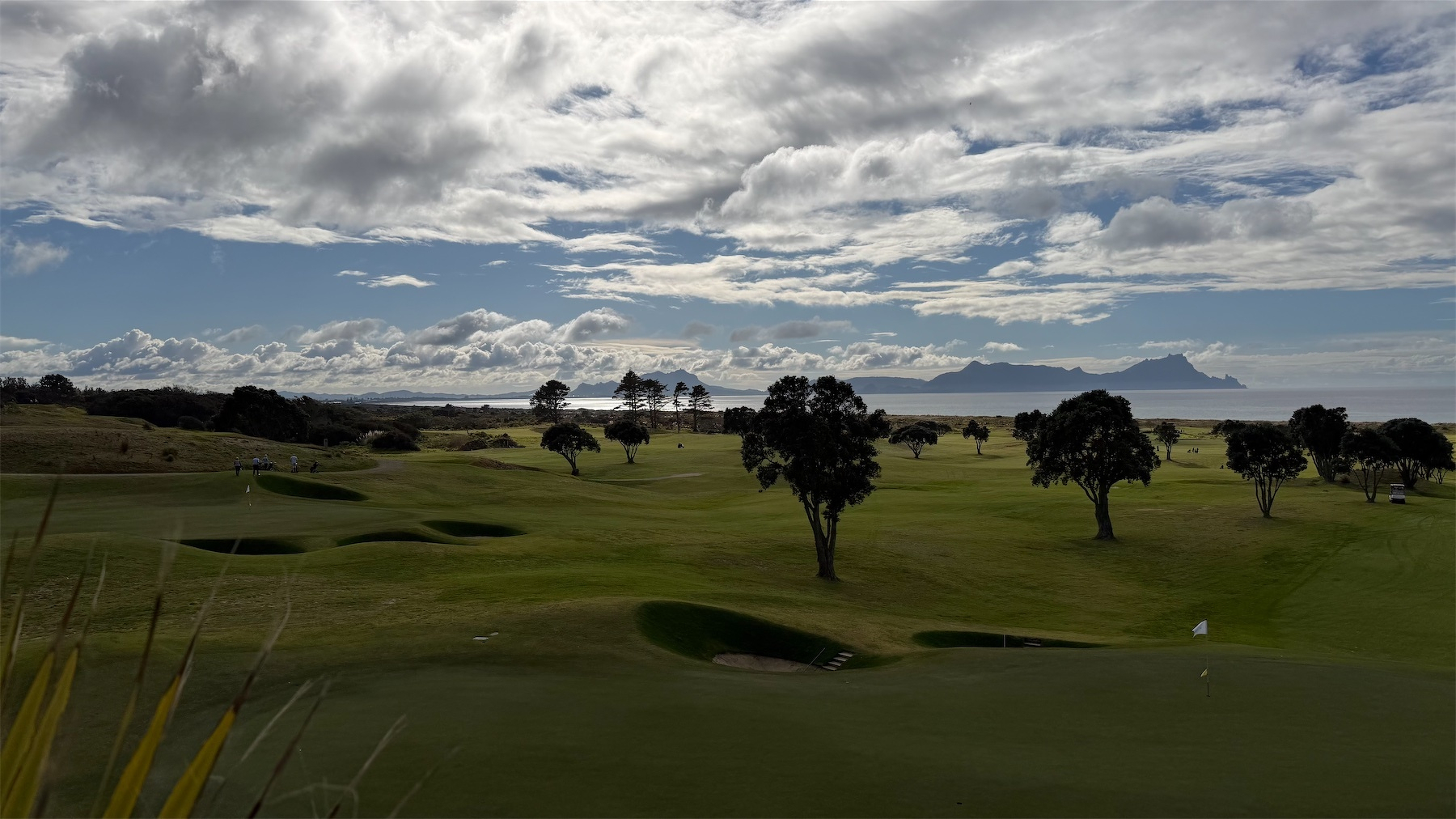 A vast golf course with scattered trees and a distant view of mountains under a partly cloudy sky. This and the following form a panorama from left to right.