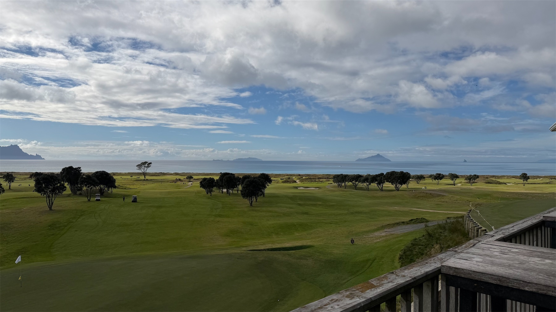 A vast, green golf course stretches out under a partly cloudy sky with distant mountains and sea visible on the horizon.
