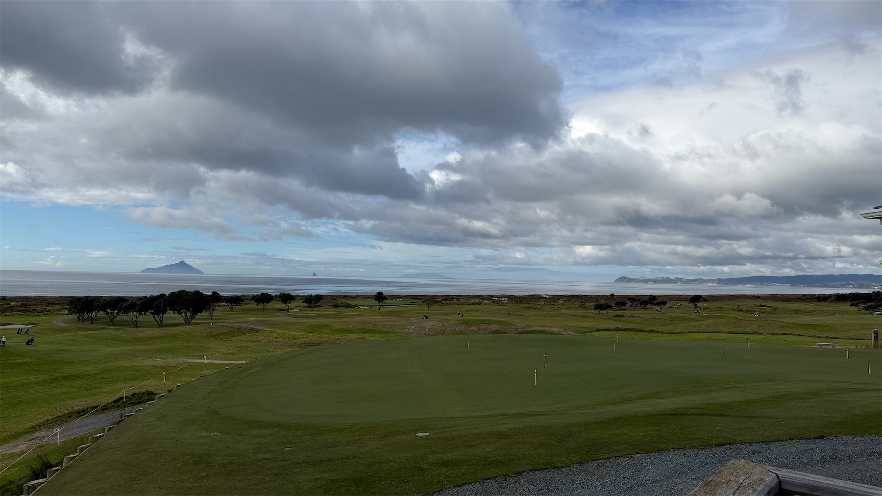 A vast golf course extends toward the ocean under a cloudy sky with distant islands visible on the horizon.