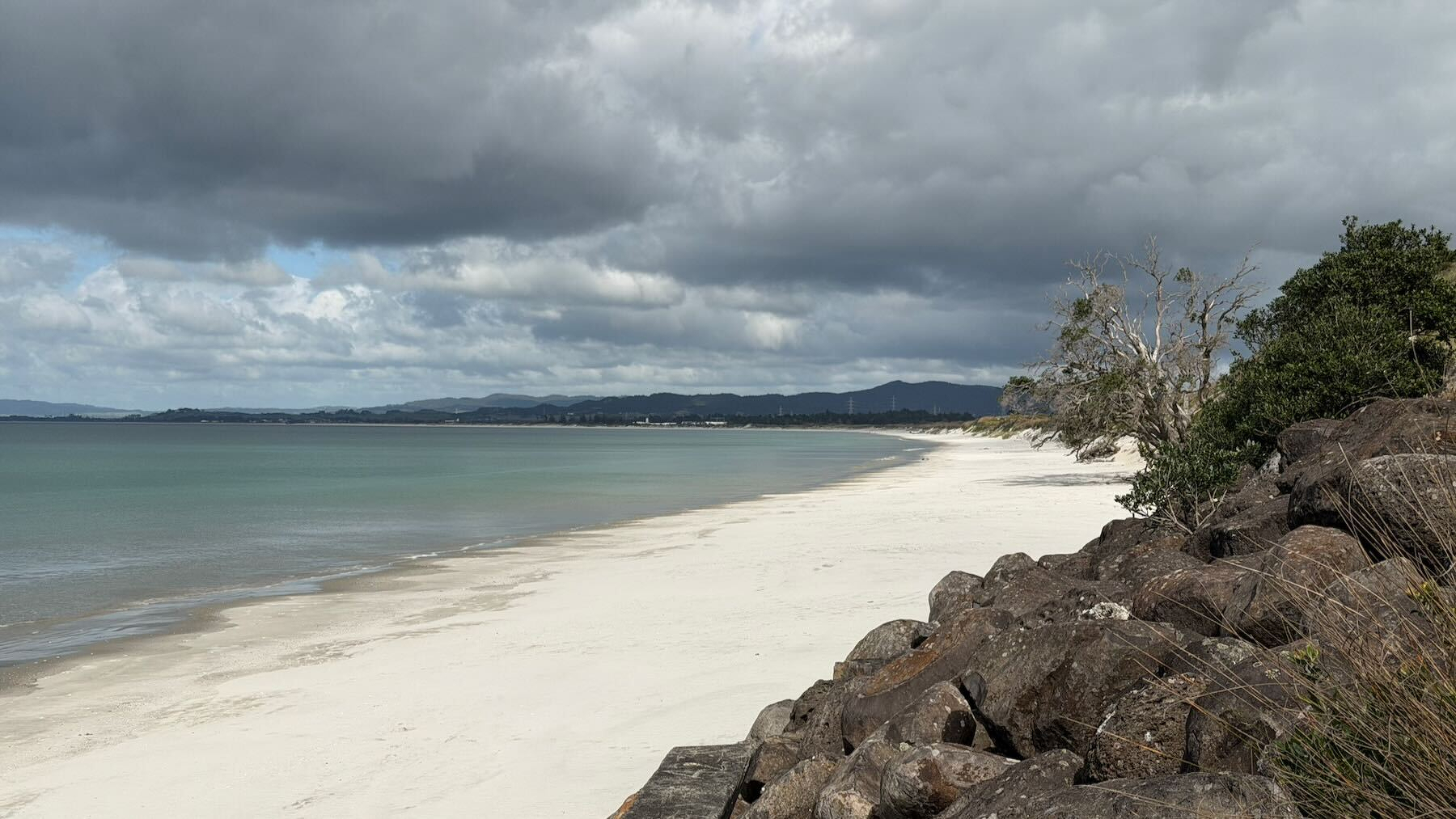 A white sandy beach stretches along the shoreline under a cloudy sky, with large rocks and some vegetation in the foreground.
