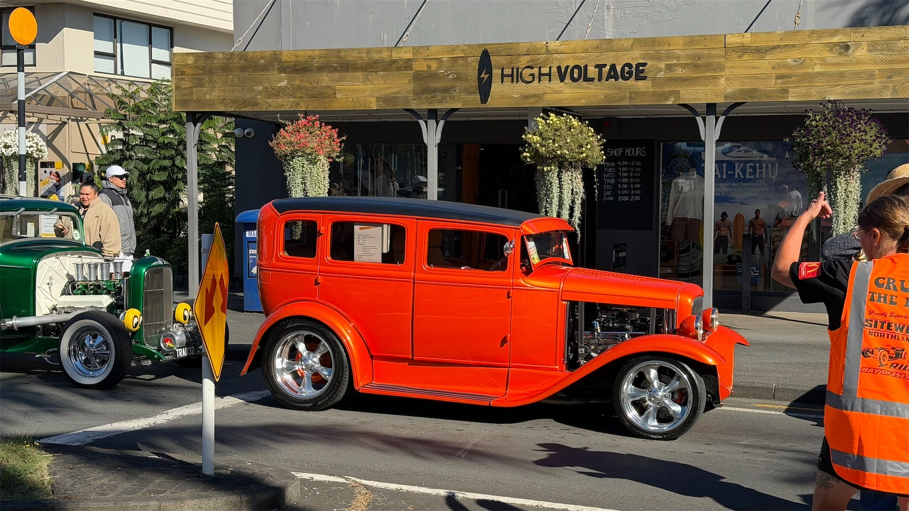 A bright orange vintage car parked in front of a building labeled High Voltage on a sunny street.