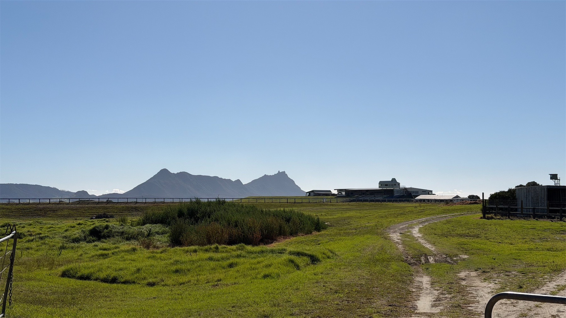 View across green grass and past the racecourse stands to hills in the distance against a clear blue sky.