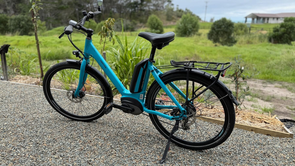 A blue bike on a stand on gravel with greenery behind. 