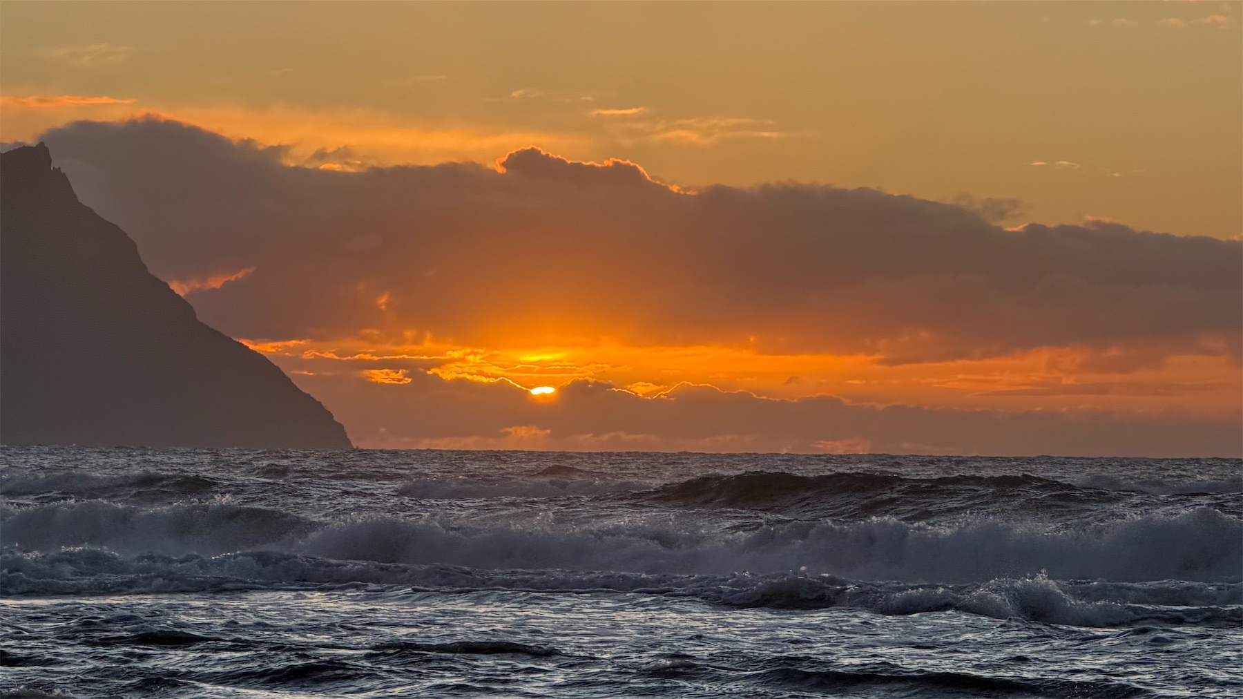 Roiling sea, a hill on the left and the top of the sun peeking through clouds on the horizon. 