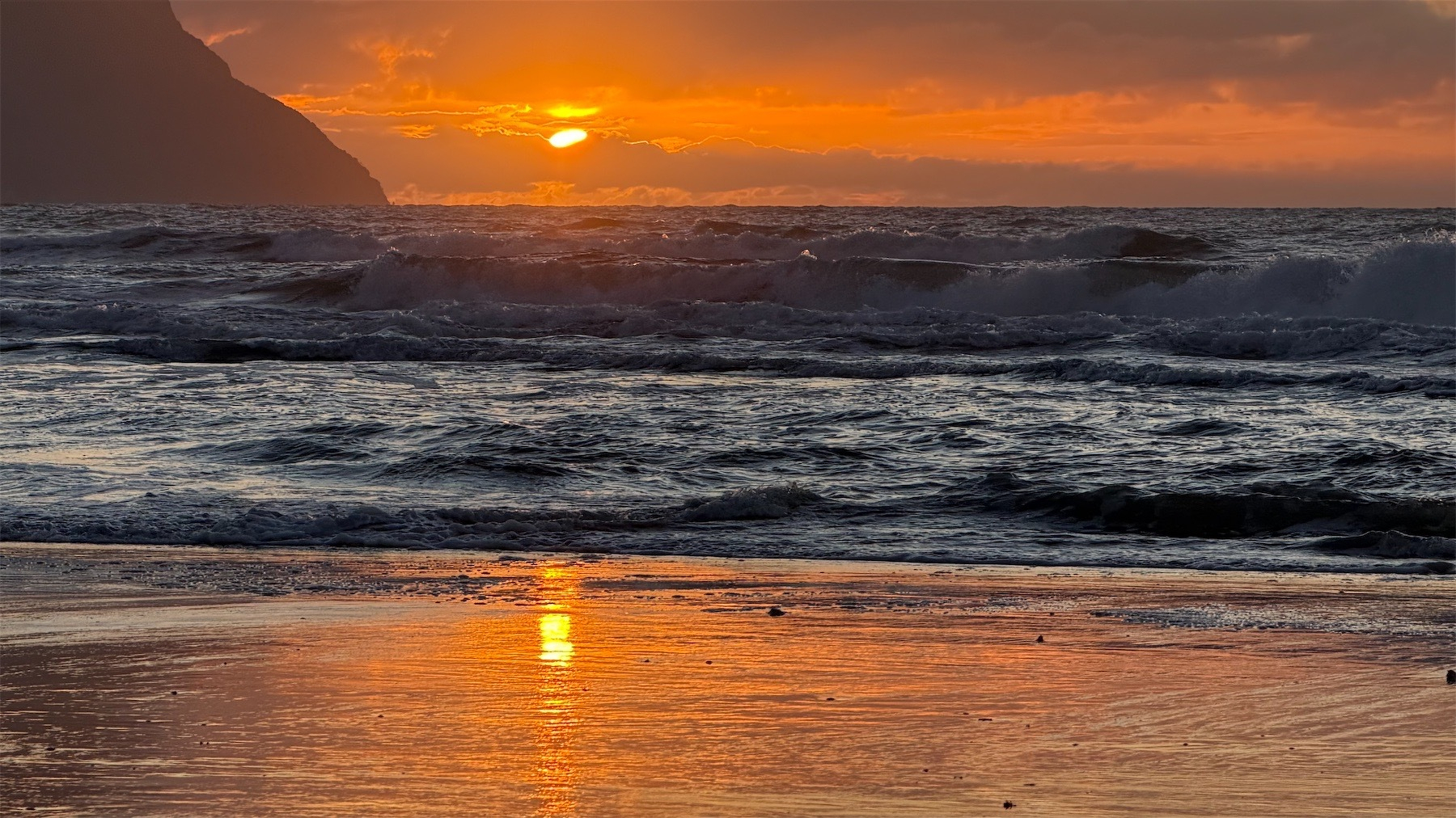 A smidgeon of sun peeking through clouds on the horizon and reflected in wet sand in front of a foaming sea; a hill on the left.