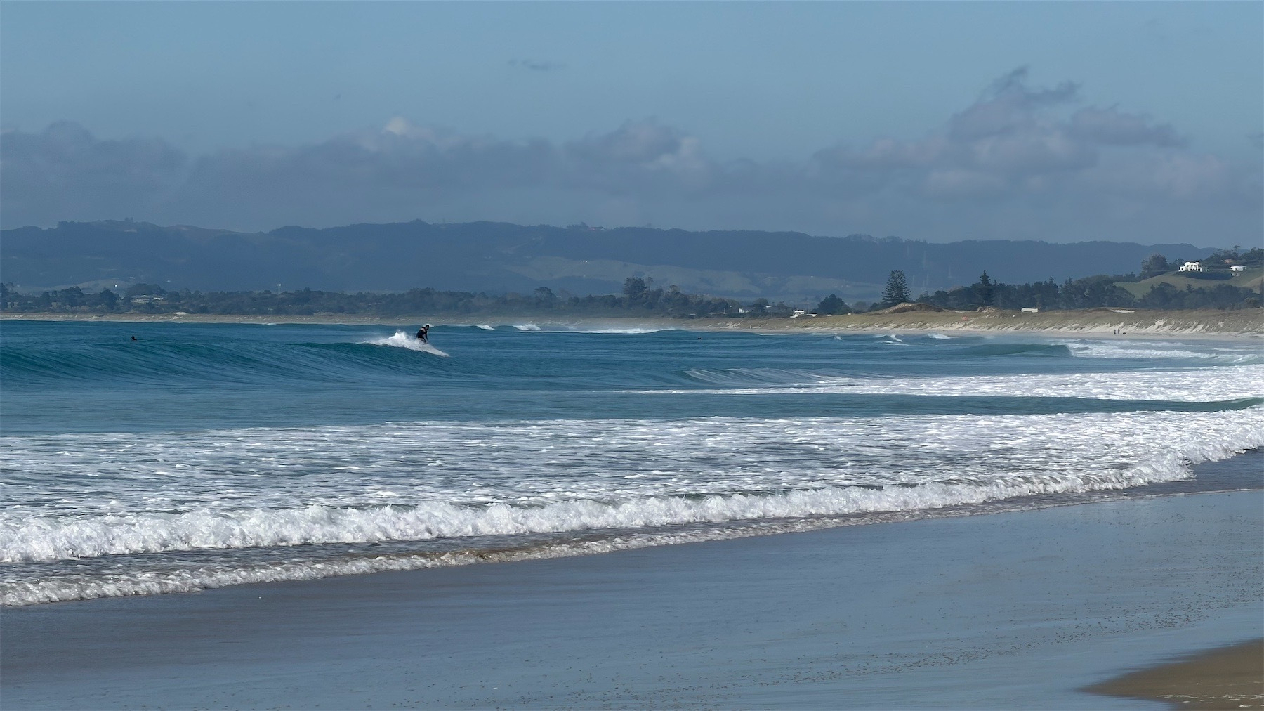 Blue sea, white water by the shore. A person is surfing a wave.