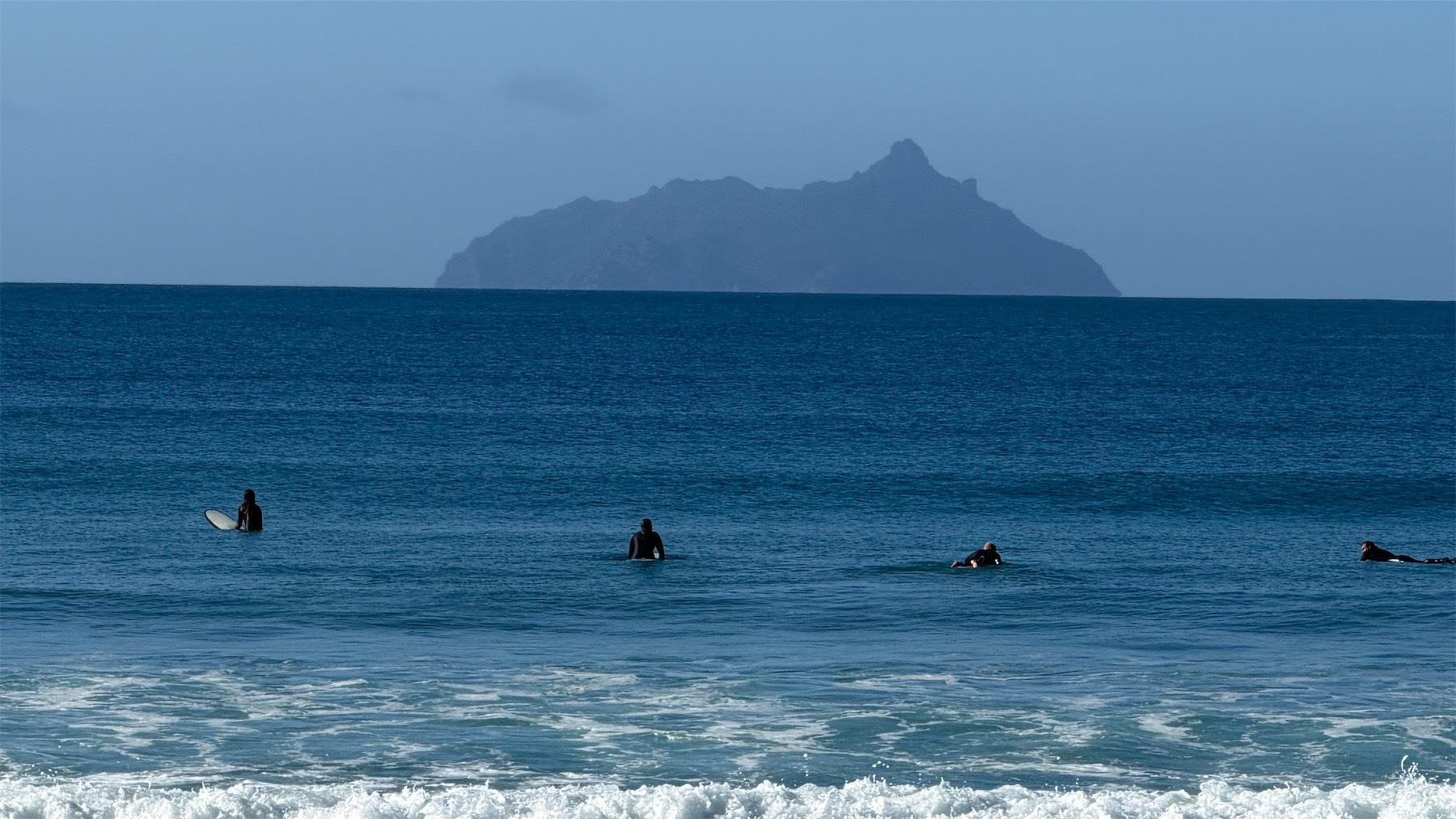 Four surfers in deep blue sea with an island behind and blue sky.