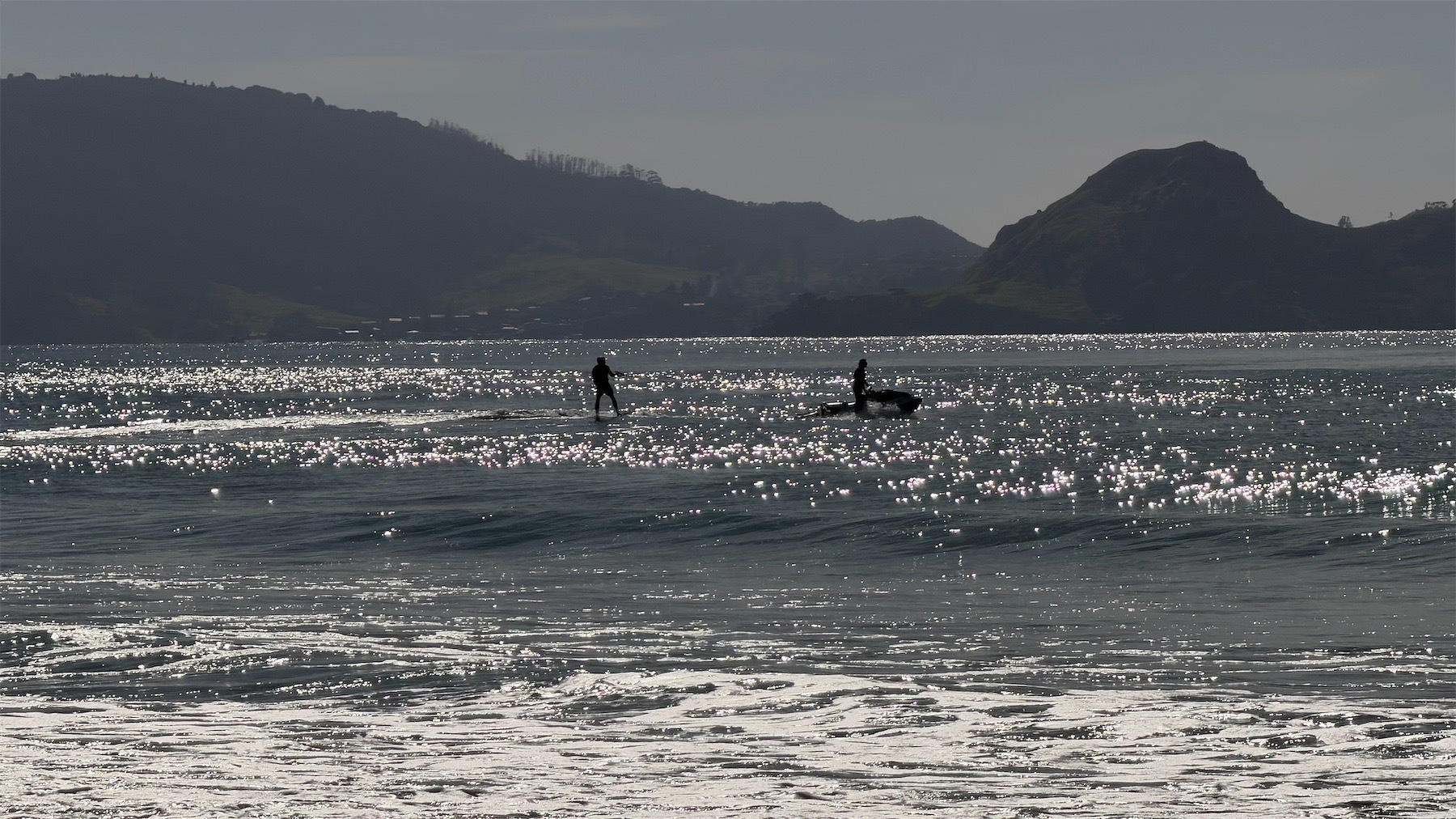 Sparkly sea with hills behind. A person is waterskiing behind a jetski.
