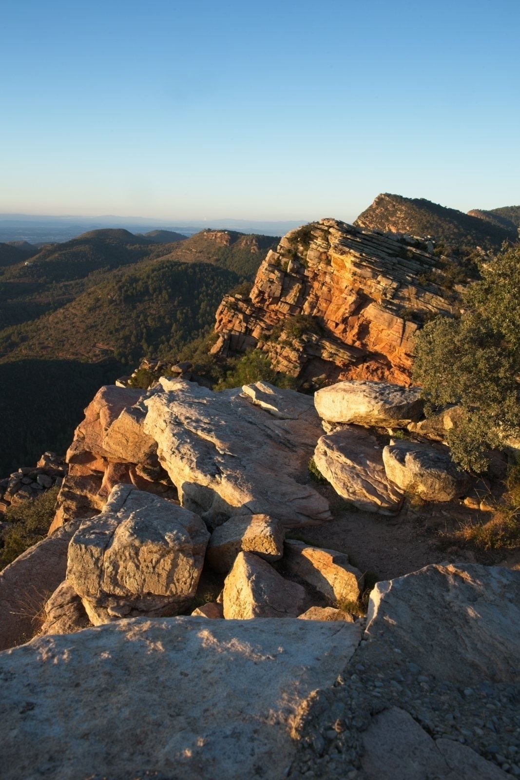 Auto-generated description: Sunlit rocky outcrop overlooks a range of distant mountains under a clear blue sky.