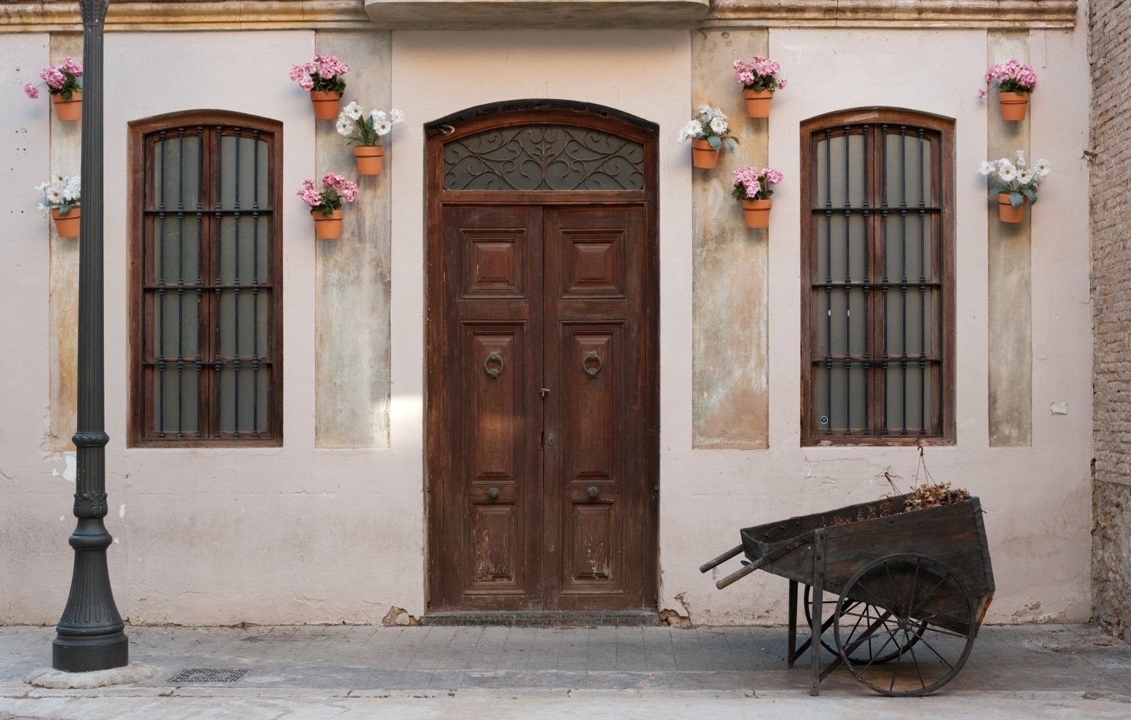 Auto-generated description: A rustic building facade features a wooden door and windows adorned with flower pots, alongside a vintage wheelbarrow in the foreground.