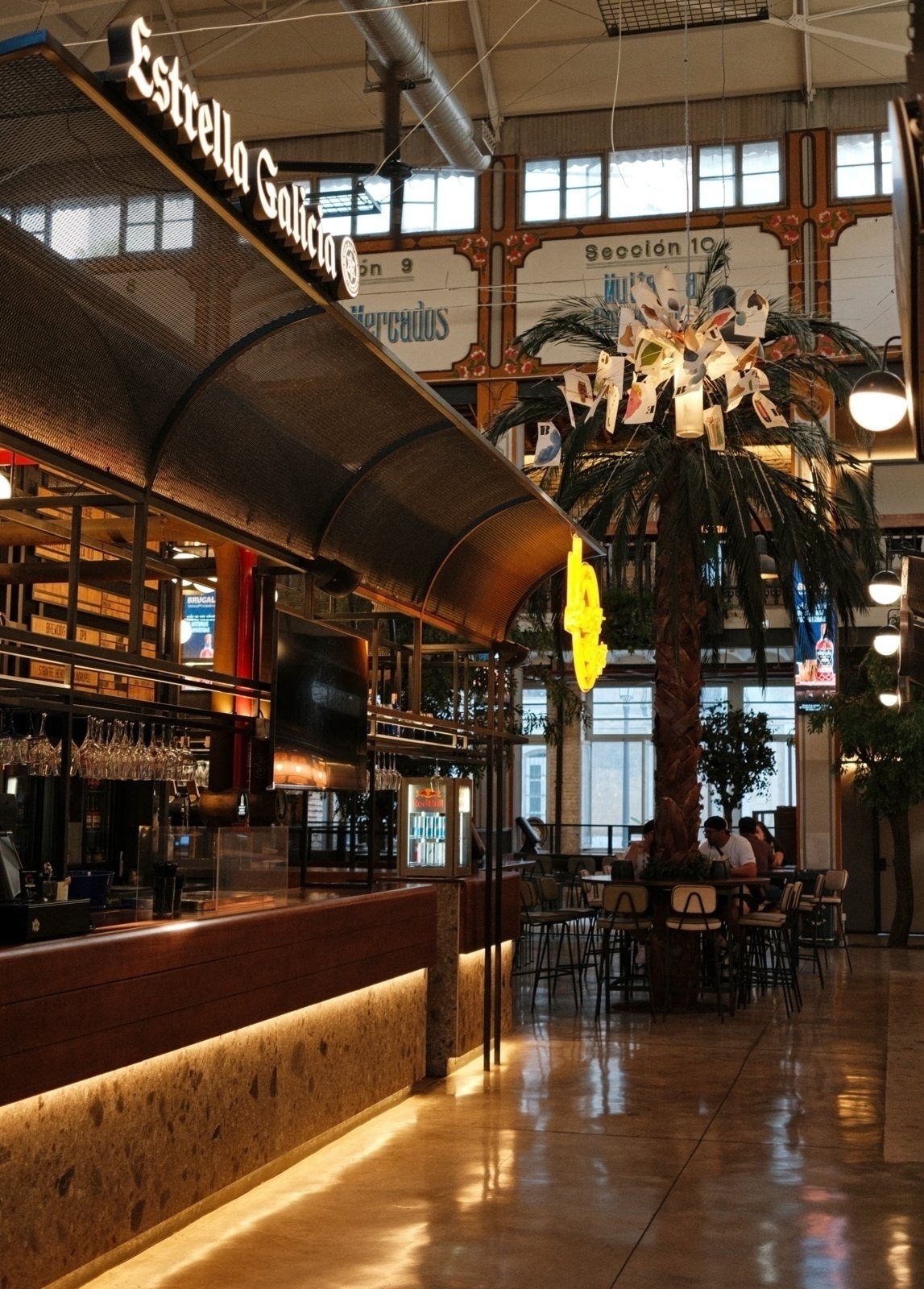 Auto-generated description: A modern indoor space features a bar with the Estrella Galicia sign, seating area, and a decorative palm tree adorned with hanging postcards.