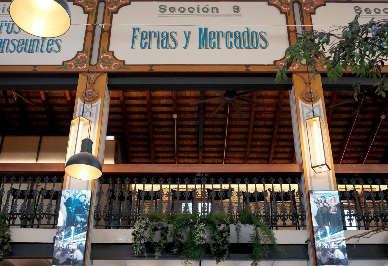 Auto-generated description: A market or fair section with a decorative sign reading Ferias y Mercados features hanging lamps, greenery, and a rustic wooden ceiling.