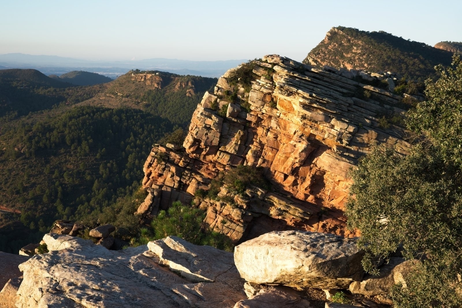 Auto-generated description: A rugged, rocky cliff overlooks a vast, hilly landscape with distant views under a clear blue sky.