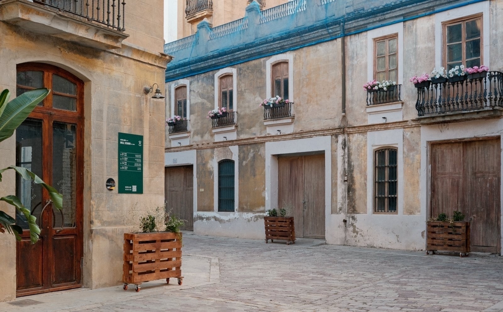 Auto-generated description: A rustic courtyard features aged buildings with wooden doors and balconies adorned with potted plants and pink flowers.