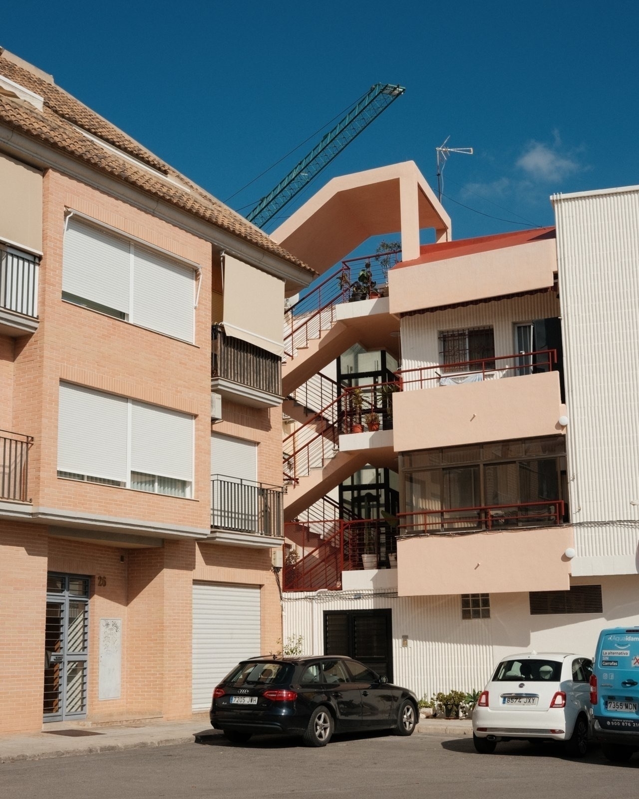 Auto-generated description: A street scene features two buildings with balconies, several parked cars, and a construction crane in the background against a clear blue sky.