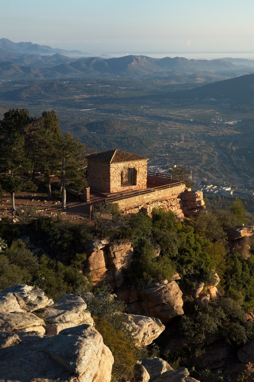 Auto-generated description: A stone building stands on a cliff surrounded by trees, overlooking a vast valley with mountain ranges in the distance.