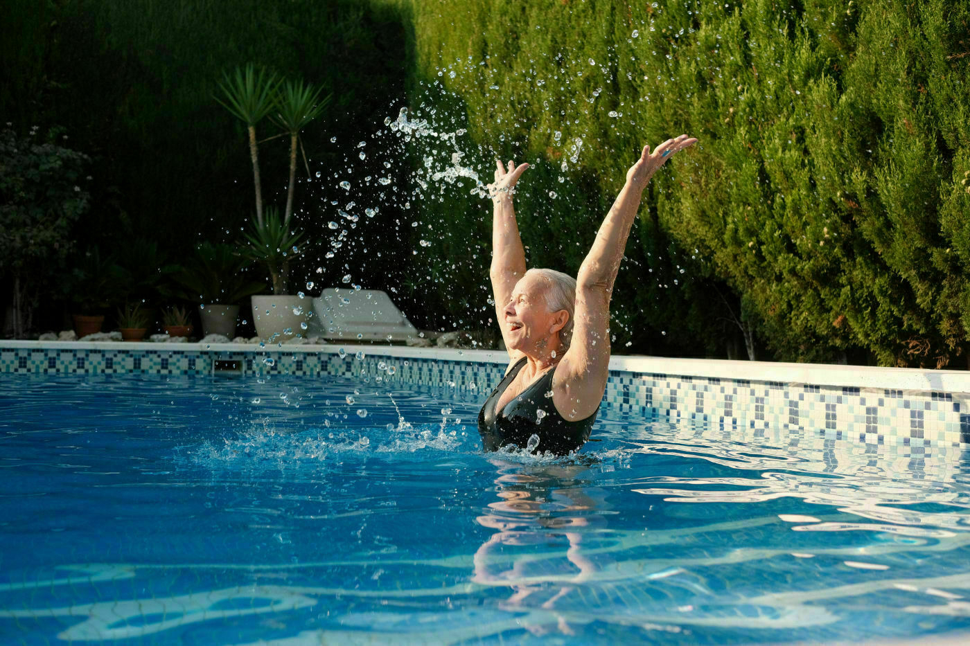 Auto-generated description: An older woman in a swimsuit joyfully raises their arms while splashing water in a swimming pool, surrounded by lush greenery.