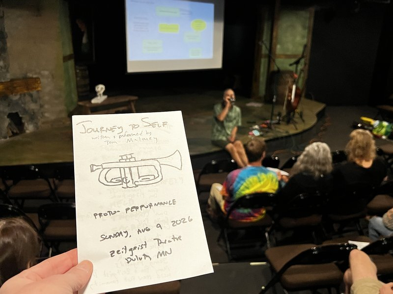 The hand-lettered program for the Journey To Self proto-performance at Zeitgeist Theatre, held up as Tom Maloney performs onstage