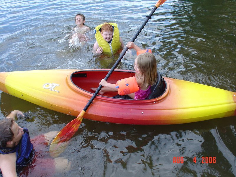 Kaylee paddling a kayak while Spencer, Corey, and a friend swim alongside in the lake