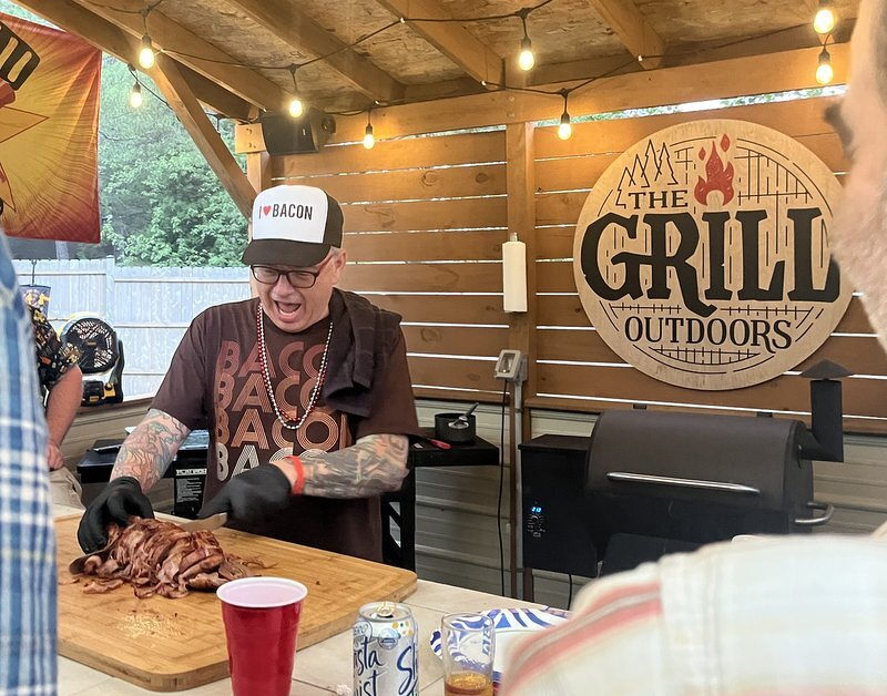 Jeff in a BACON hat carving one of his smoked meat logs under the string lights of the backyard grilling pavilion