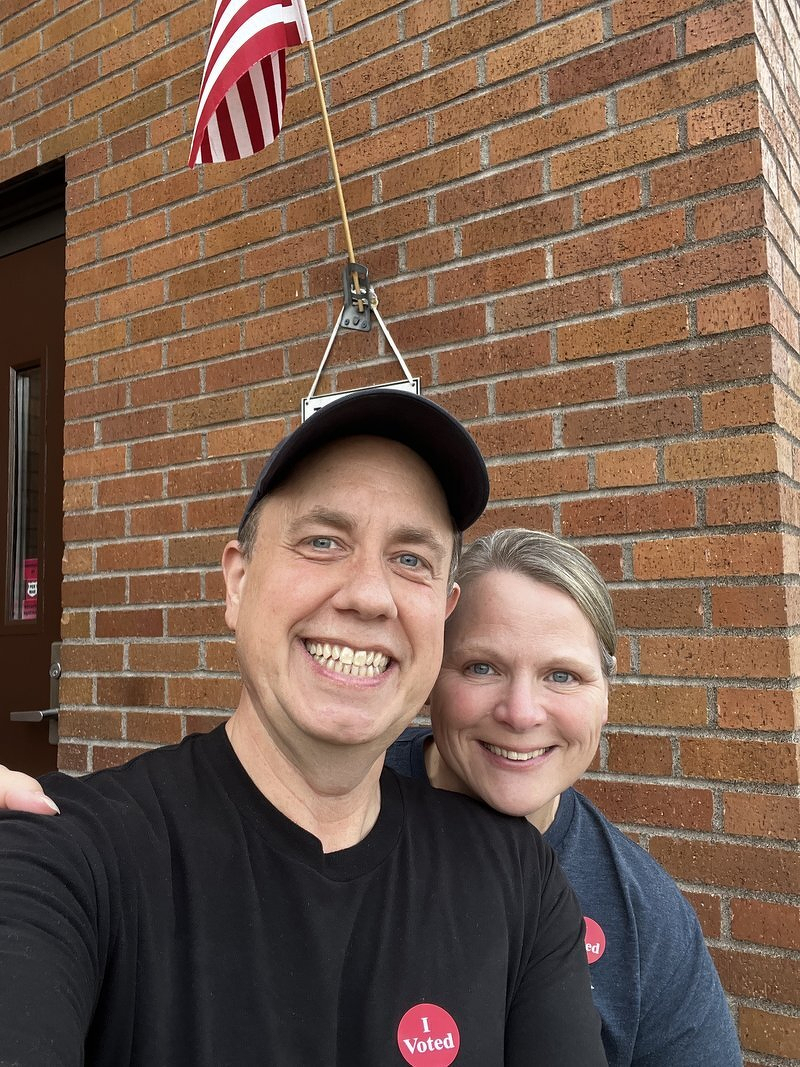 Justin and Jody with 'I Voted' stickers outside the polling place, an American flag on the brick wall behind them