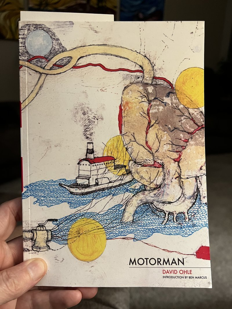 A copy of Motorman by David Ohle held up, its cover a surreal drawing of a tugboat, a giant hand, and a yellow sun