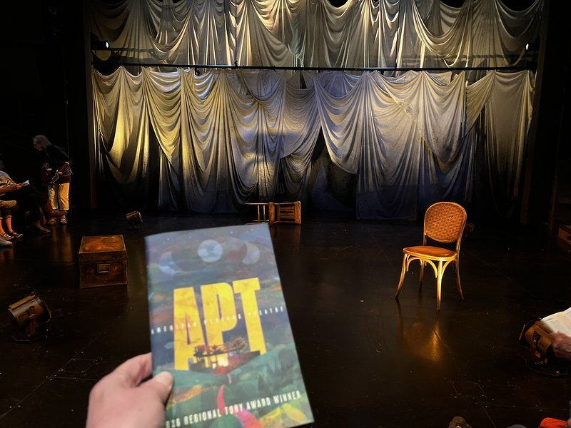 The draped set of The Chairs in the Touchstone Theatre with a couple of empty chairs, an APT program held up in the foreground