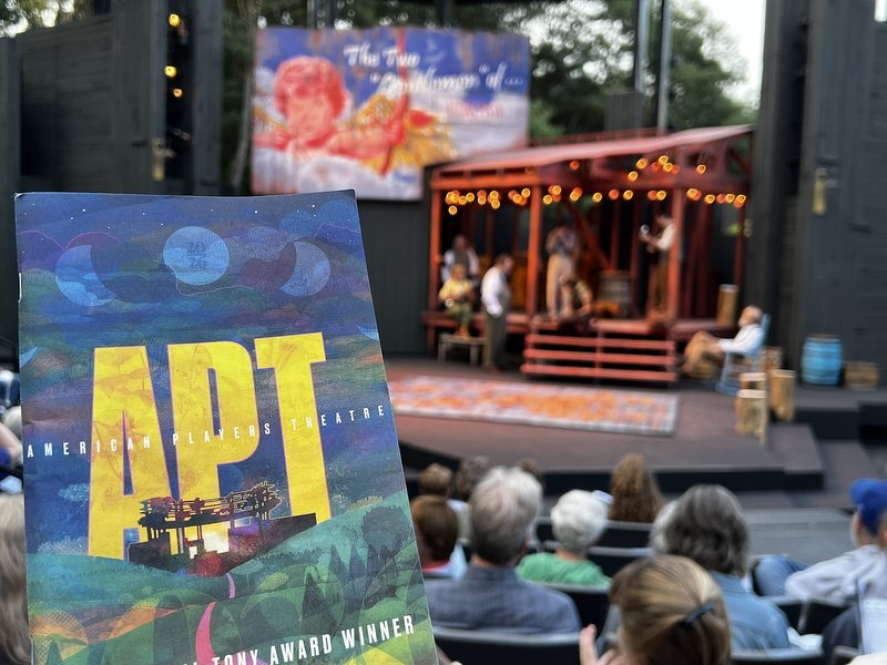 The Two Gentlemen of Verona title projected above the red bandstand set in the Hill Theatre, an APT program held up in the foreground