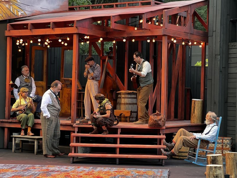 The cast of The Two Gentlemen of Verona on the string-lit porch set, one actor at the microphone