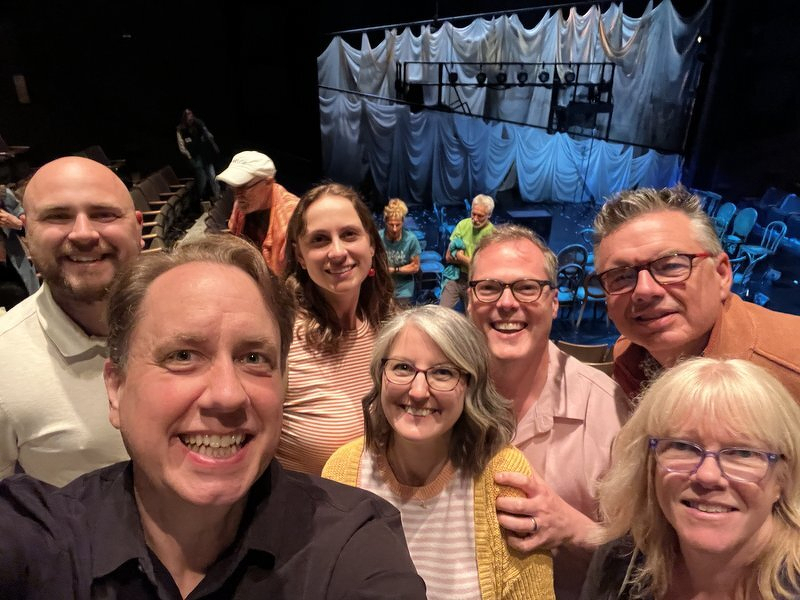 The group crowded together for a selfie inside the Touchstone Theatre, the draped set behind them