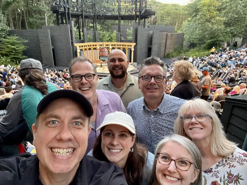 The group smiling in the Hill Theatre seats at dusk, the wooden set glowing on stage