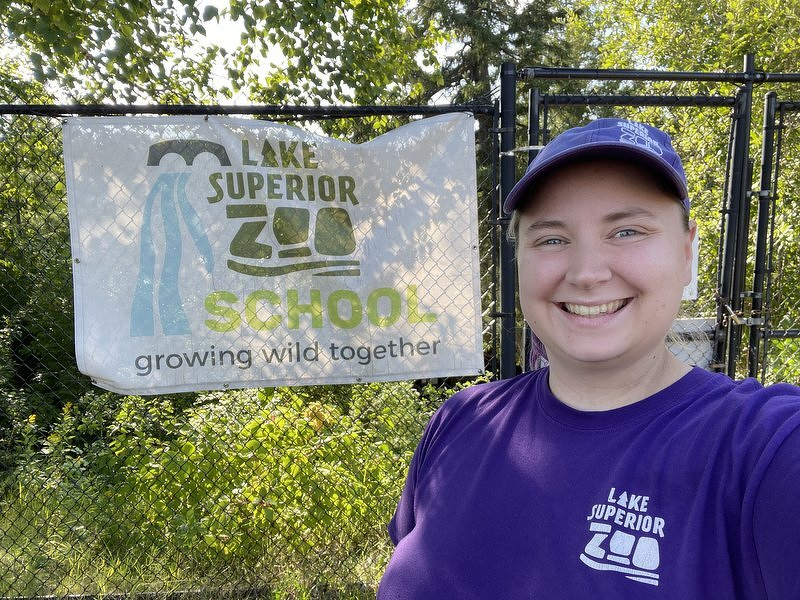 Kaylee in a Lake Superior Zoo t-shirt and ballcap, smiling in front of the Lake Superior Zoo School banner