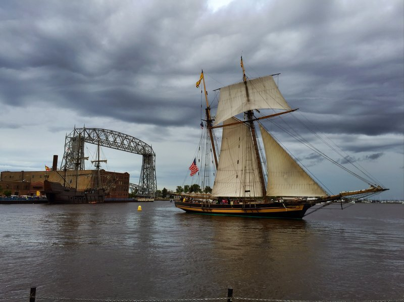 A tall ship under sail passing the Aerial Lift Bridge in the Duluth harbor under storm clouds