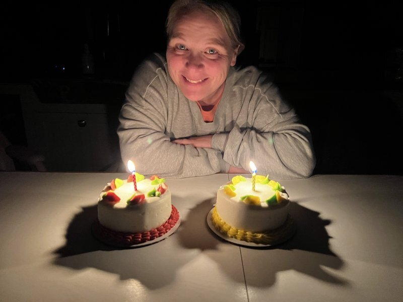 Jody smiling behind two small birthday cakes lit with candles in a dark kitchen