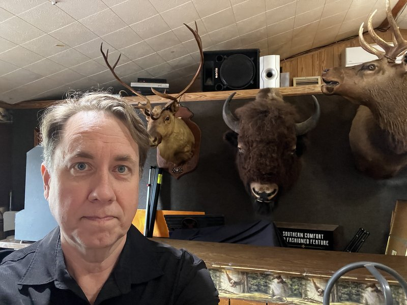 Justin taking a deadpan selfie in front of a wall of mounted bison, elk, and deer heads