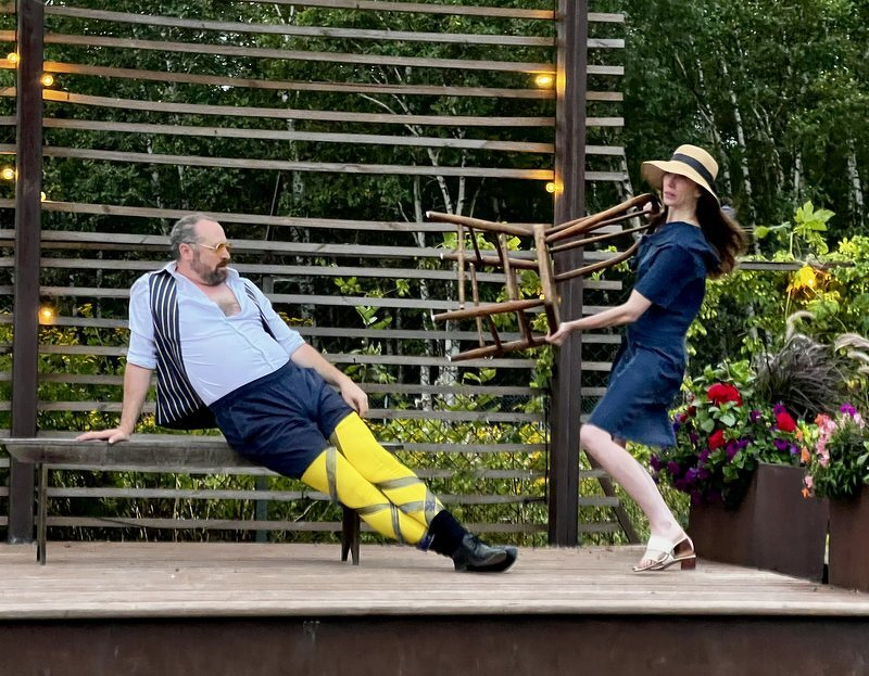Malvolio in yellow stockings sprawled on a bench while Maria hauls a chair away, on the outdoor stage at Castle Danger Brewery