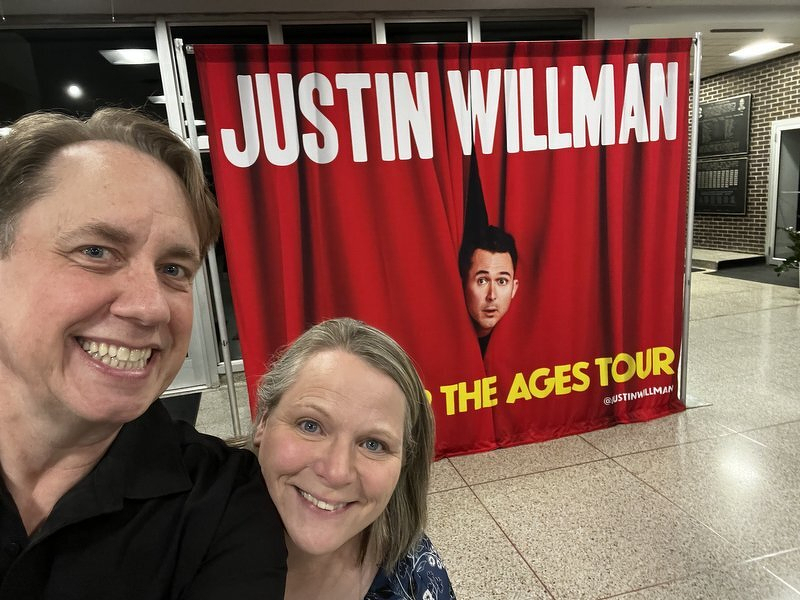 Justin and Jody grinning for a selfie in front of the red Justin Willman: The Ages Tour banner