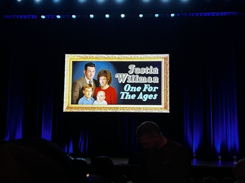 The blue-lit DECC stage with the gilt-framed Justin Willman: One For The Ages title card