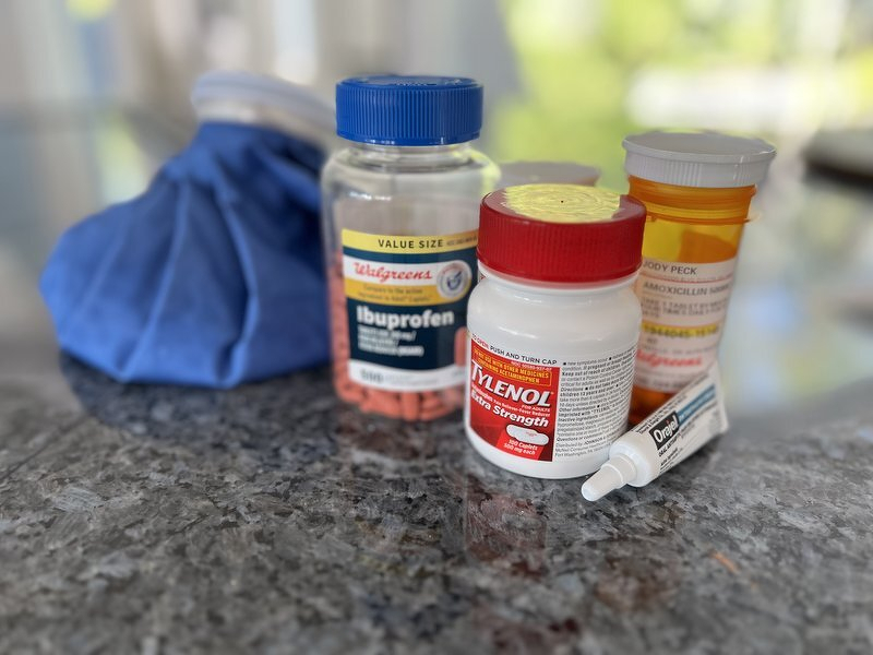 A cold pack, bottles of ibuprofen and Tylenol, and a tube of Orajel lined up on the kitchen counter