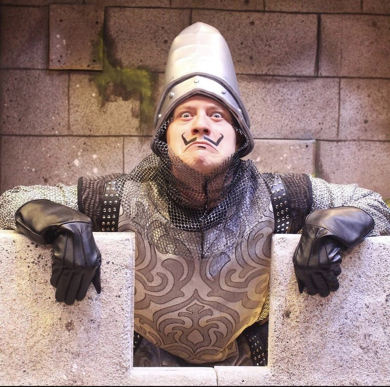 Phil as a French taunter in a tall helmet, chainmail, and curled mustache, leaning over the castle wall with gloved hands dangling