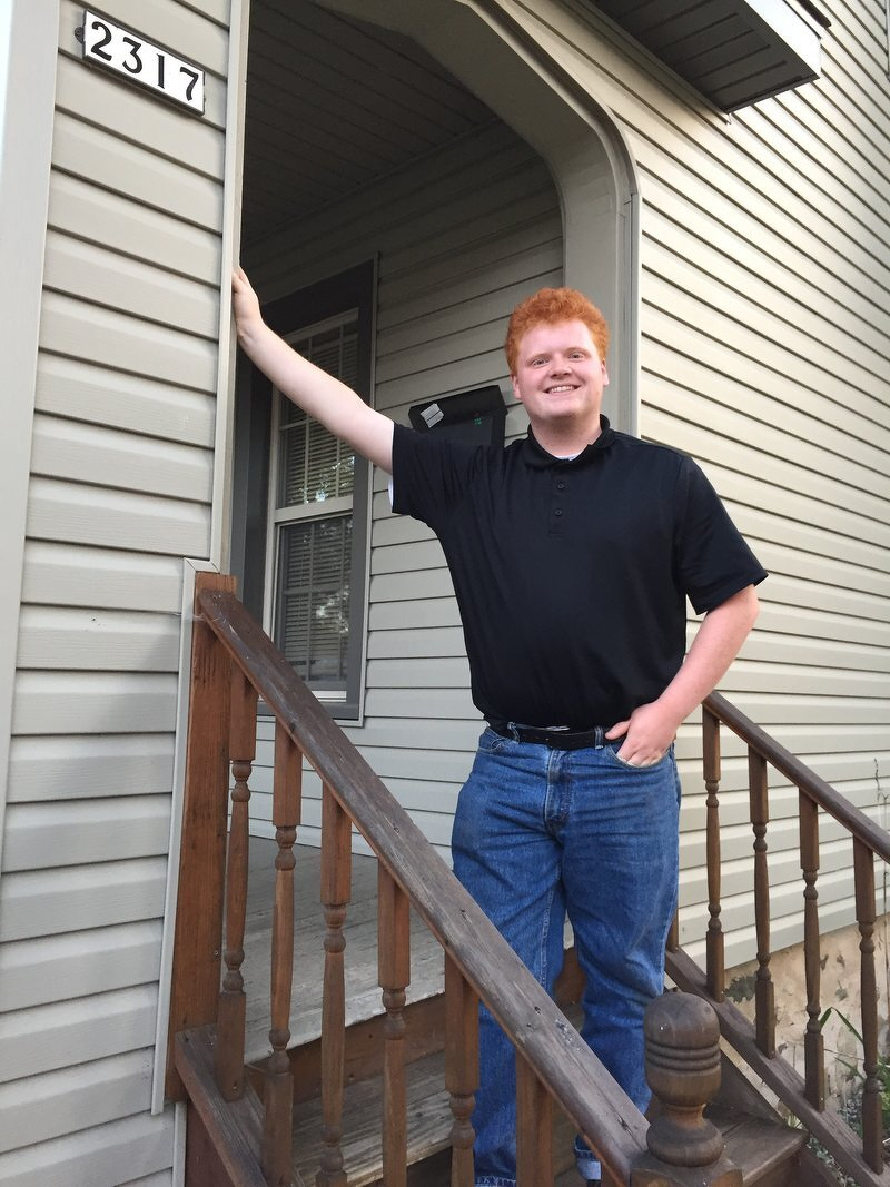 Spencer in a black polo and jeans, smiling on the front steps of the Five Guys house