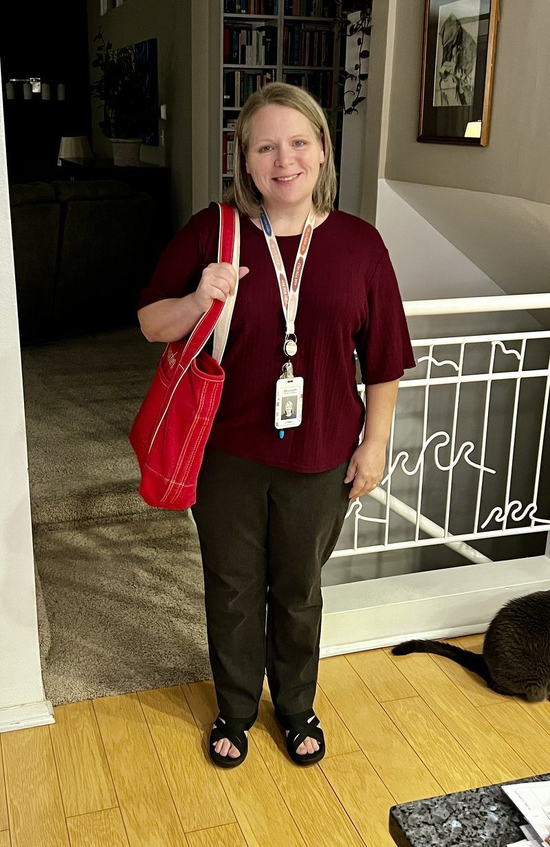 Jody smiling in the entryway on the first day of school, wearing a maroon top and a lanyard, with a red tote bag over her shoulder