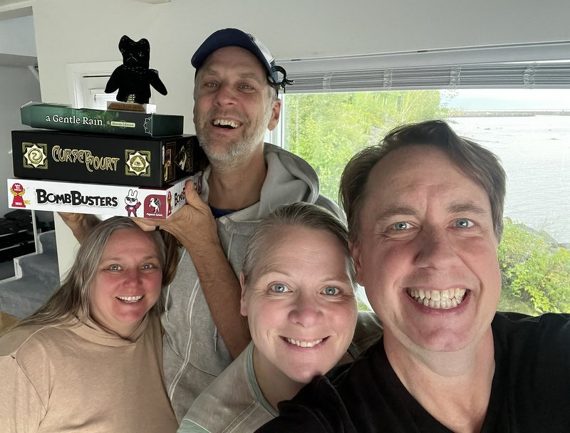 Tom, Brandy, Jody, and Justin grinning in a selfie with a stack of board games, including Bomb Busters, and the lake behind them